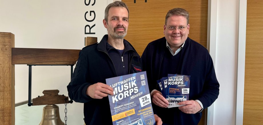 Konzertankündigung Zwei Männer stehen in einem Innenraum neben einer großen, freihängenden Glocke aus Metall in einem Holzgestell. Beide halten Plakate bzw. Flyer mit der Aufschrift „Luftwaffen Musikkorps Münster“. Auf dem Plakat steht unter anderem „Charity Konzert“ sowie das Datum „28. April“; weitere Angaben zu Ort und Uhrzeit sind abgedruckt. Im Hintergrund sind eine Holztür und eine Wand mit teilweise sichtbarer Beschriftung zu sehen.