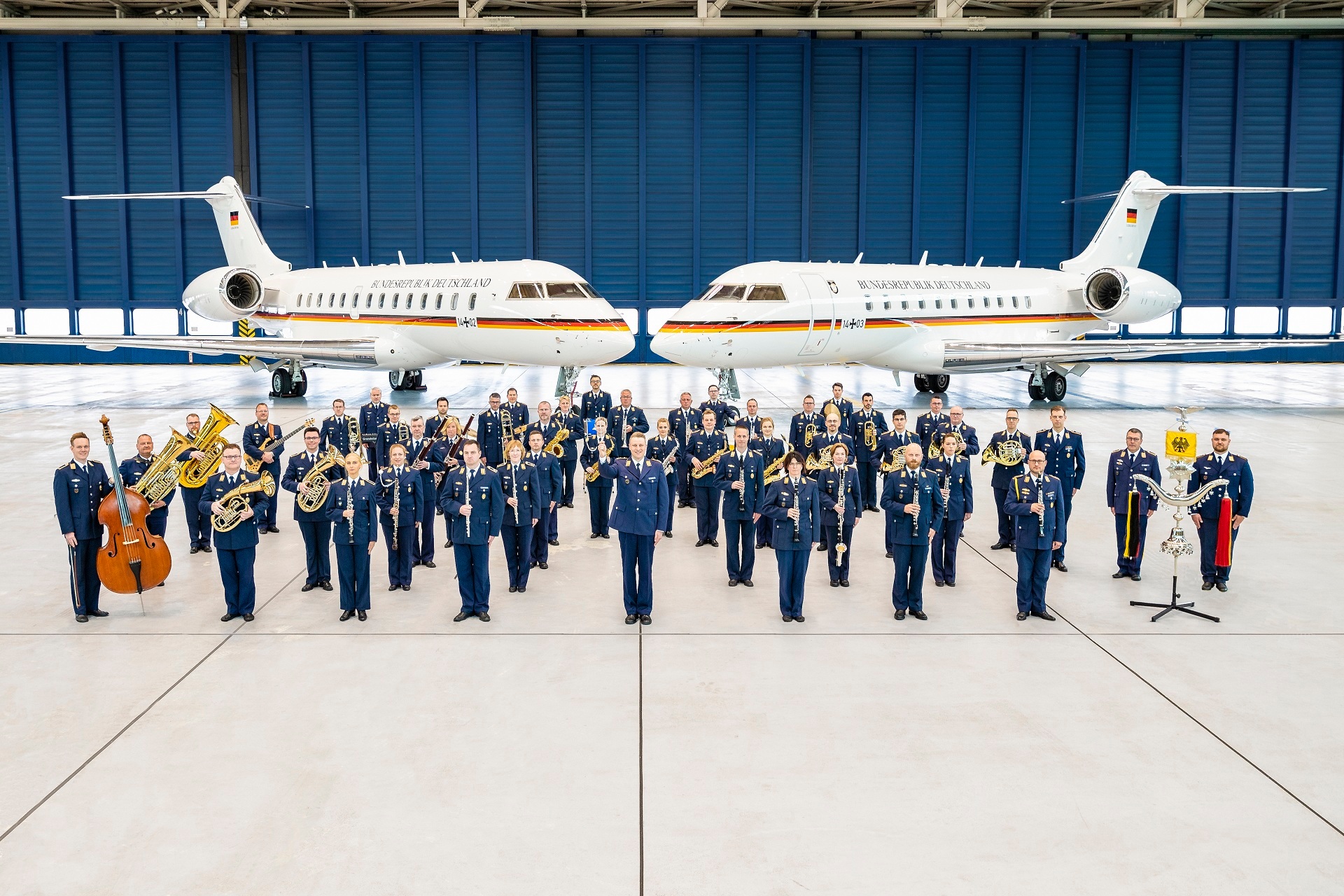 Rund 50 Personen eines Orchesters stehen in blauen Uniformen der Luftwaffe und mit verschiedenen Instrumenten in einem Flugzeughangar. Im Hintergrund stehen zwei Flugzeuge der Luftwaffe.