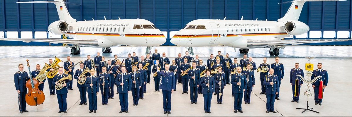 Luftwaffenmusikkorps der Luftwaffe vor Global 5000 der FlgBschft. Rund 50 Personen eines Orchesters stehen in blauen Uniformen der Luftwaffe und mit verschiedenen Instrumenten in einem Flugzeughangar. Im Hintergrund stehen zwei Flugzeuge der Luftwaffe.