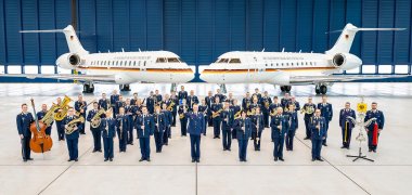 Rund 50 Personen eines Orchesters stehen in blauen Uniformen der Luftwaffe und mit verschiedenen Instrumenten in einem Flugzeughangar. Im Hintergrund stehen zwei Flugzeuge der Luftwaffe.