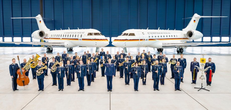 Rund 50 Personen eines Orchesters stehen in blauen Uniformen der Luftwaffe und mit verschiedenen Instrumenten in einem Flugzeughangar. Im Hintergrund stehen zwei Flugzeuge der Luftwaffe.
