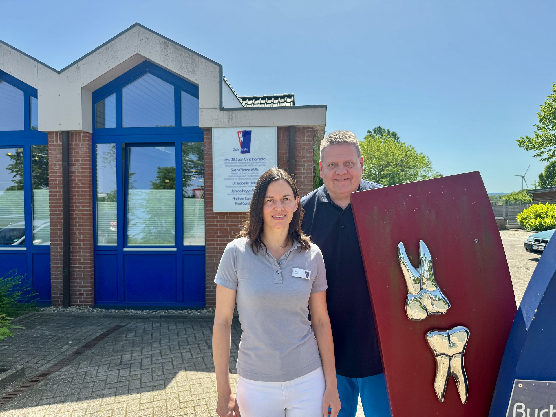 Zwei Personen stehen vor einer Zahnarztpraxis an einem sonnigen Tag. Die Frau trägt ein hellgraues Poloshirt und weiße Hose, der Mann ein dunkles Polohemd. Neben ihnen steht eine große rote Tafel mit zwei metallischen Zahnmodellen. Im Hintergrund sind das Praxisgebäude mit blauen Fensterrahmen, ein Praxisschild sowie Bäume und blauer Himmel zu sehen.