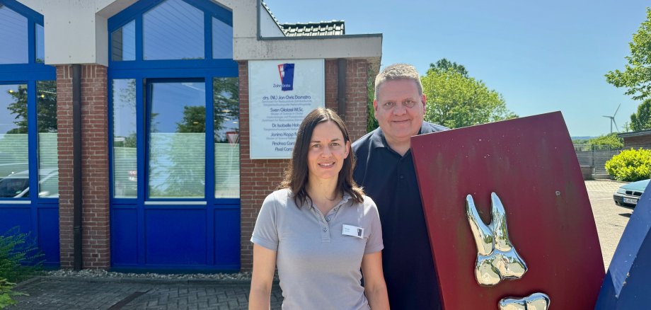 Zwei Personen stehen vor einer Zahnarztpraxis an einem sonnigen Tag. Die Frau trägt ein hellgraues Poloshirt und weiße Hose, der Mann ein dunkles Polohemd. Neben ihnen steht eine große rote Tafel mit zwei metallischen Zahnmodellen. Im Hintergrund sind das Praxisgebäude mit blauen Fensterrahmen, ein Praxisschild sowie Bäume und blauer Himmel zu sehen.