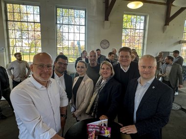 Gruppenfoto mit zwei Frauen und fünf Männern in einem Veranstaltungsraum, der wirkt wie eine Industriehalle.