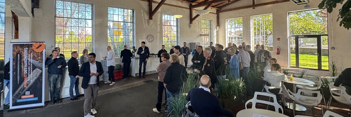 Großer, heller Veranstaltungsraum mit hohen Holzdecken und großen Fenstern. Mehrere Menschen stehen und sitzen in kleinen Gruppen bei einem Networking- oder Informations­treffen. Im Vordergrund befinden sich runde Tische und weiße Stühle, rechts sitzen einige Personen. Tageslicht fällt durch die Fenster und beleuchtet den Raum.