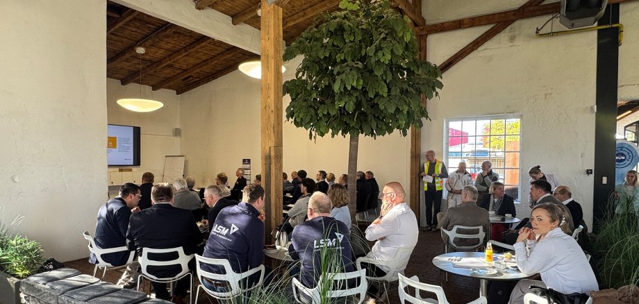 Veranstaltung in einem großen Saal mit hoher Holzdecke und großen Fenstern. Zahlreiche Menschen sitzen an Tischen oder auf Stühlen und verfolgen einen Vortrag. Vorne stehen zwei Referenten neben einem Bildschirm mit Präsentationsfolie. In der Mitte des Raums steht ein großer grüner Innenbaum. Tageslicht erhellt den Raum, einige Teilnehmende stehen am Rand der Veranstaltung.