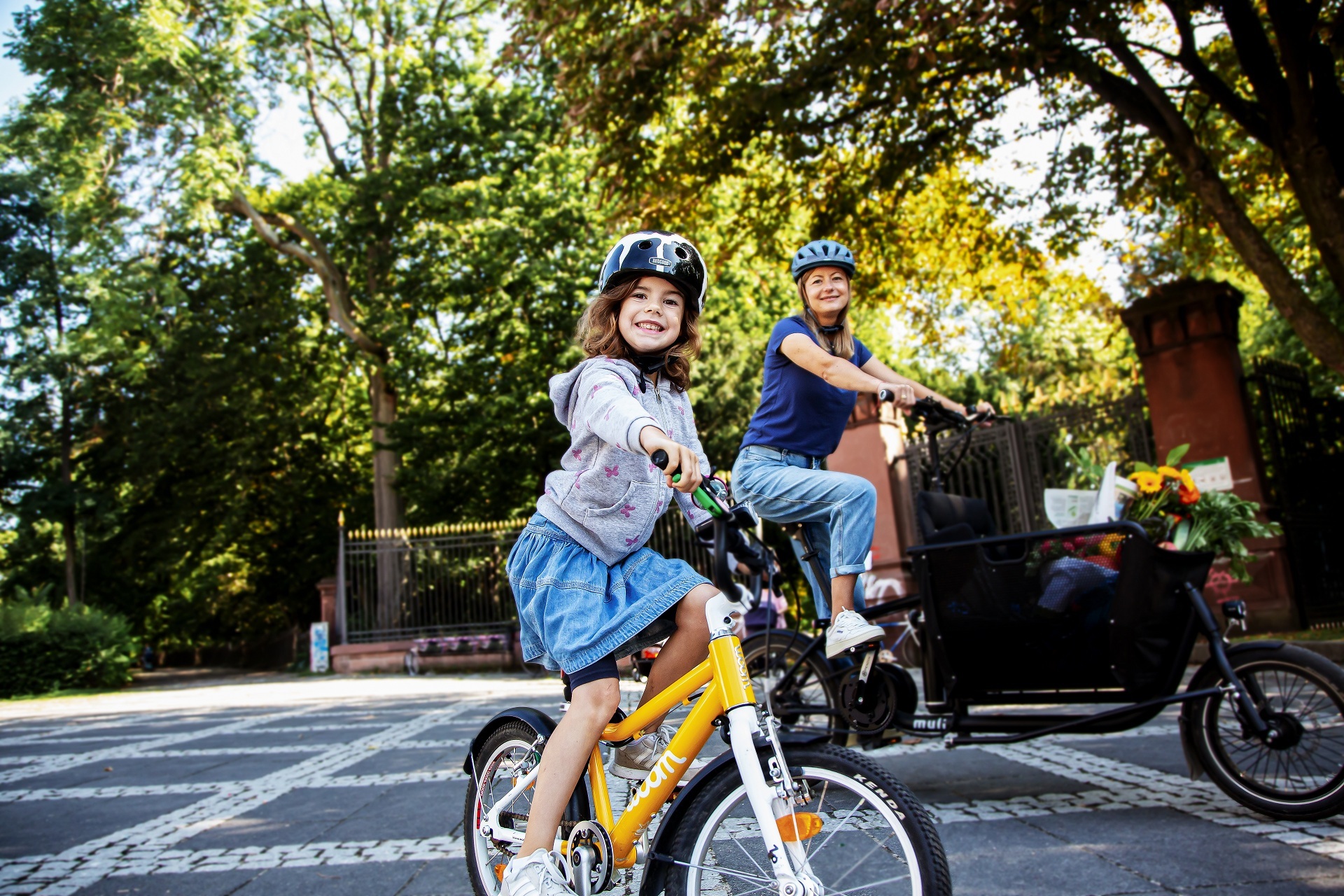 Ein Kind mit Fahrradhelm fährt auf einem gelben Kinderfahrrad im Vordergrund und lächelt in die Kamera. Dahinter fährt eine erwachsene Person mit Helm auf einem Lastenfahrrad. Beide befinden sich auf einer ruhigen, von Bäumen gesäumten Straße bei sonnigem Wetter. Im Lastenrad sind Taschen und Blumen zu sehen.