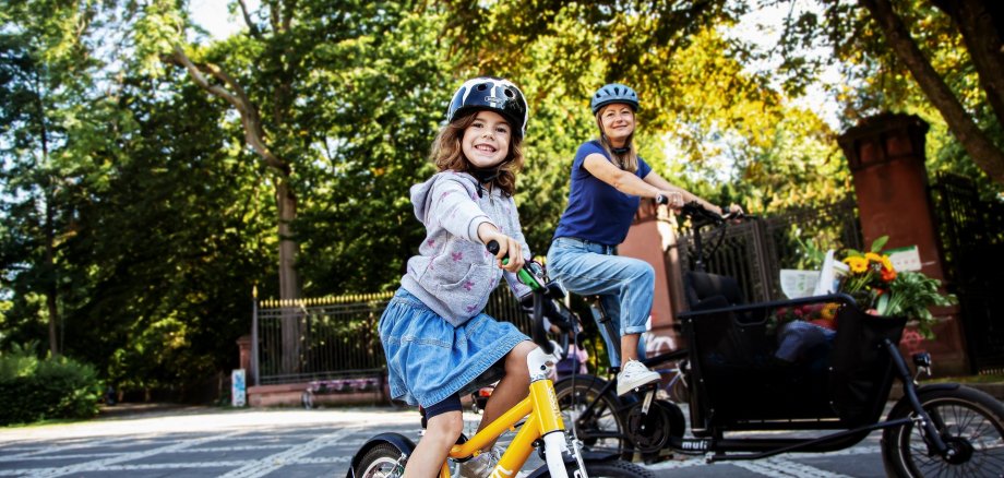 STADTRADELN Ein Kind mit Fahrradhelm fährt auf einem gelben Kinderfahrrad im Vordergrund und lächelt in die Kamera. Dahinter fährt eine erwachsene Person mit Helm auf einem Lastenfahrrad. Beide befinden sich auf einer ruhigen, von Bäumen gesäumten Straße bei sonnigem Wetter. Im Lastenrad sind Taschen und Blumen zu sehen.