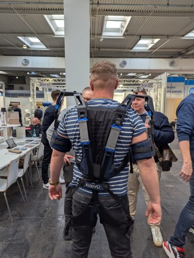 Innovationspfad 2026 Ein Mann steht auf einer Fachmesse von hinten im Mittelpunkt. Er trägt über einem blau-weiß gestreiften T-Shirt ein schwarzes Exoskelett mit gepolsterten Schulter- und Hüftgurten. Mehrere Personen stehen um ihn herum und beobachten die Vorführung. Im Hintergrund sind Messestände und ein Hallendach mit Oberlichtern zu sehen.