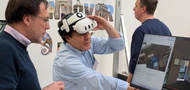 Innovationspfad 2026 Drei Männer an einem Messestand für digitale Planung: Einer trägt eine Virtual-Reality-Brille und bedient einen Computerbildschirm, auf dem ein digitaler Gebäuderaum zu sehen ist. Ein weiterer Mann beobachtet ihn, während ein dritter im Hintergrund steht.