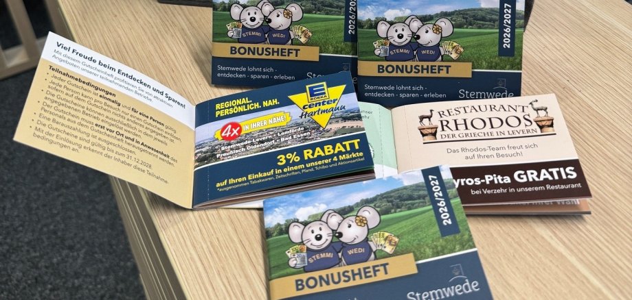 Bonusheft 2026 Mehrere Exemplare des Stemweder Bonushefts liegen auf einem Tisch in der Tourist-Info. Auf dem Titel sind die Maskottchen Stemmi und Wedi vor einer Landschaft zu sehen. Im Hintergrund stehen zwei Stoffmäuse mit Stemwede-Schals in einer dekorierten Holzkiste.