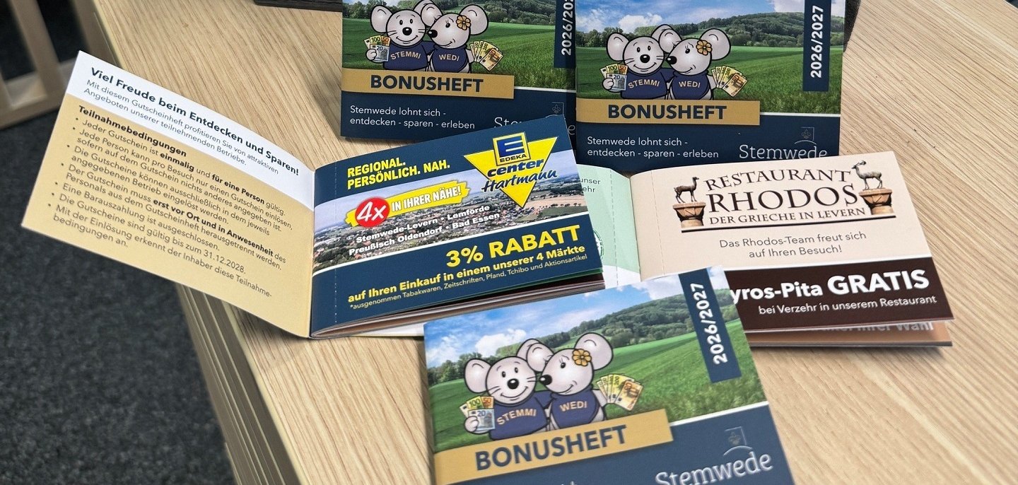 Mehrere Exemplare des Stemweder Bonushefts liegen auf einem Tisch in der Tourist-Info. Auf dem Titel sind die Maskottchen Stemmi und Wedi vor einer Landschaft zu sehen. Im Hintergrund stehen zwei Stoffmäuse mit Stemwede-Schals in einer dekorierten Holzkiste.