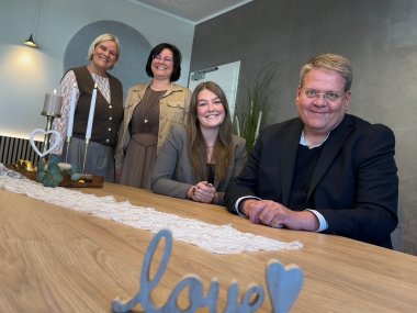 Vier Personen sitzen und stehen lächelnd an einem hellen Holztisch in einem wohnlich eingerichteten Raum. Im Vordergrund steht eine Dekoration mit dem Schriftzug „Love“ und einem Herz. Auf dem Tisch befinden sich außerdem Kerzen und ein weißer Tischläufer.