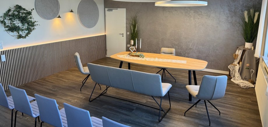 Modern eingerichteter Trau- und Veranstaltungsraum mit großem Holztisch, grauen Stühlen und Sitzbank. Über dem Tisch hängen zwei runde Leuchten; Kerzen und florale Dekoration schmücken die Tischmitte. An den Wänden befinden sich Pflanzen, runde Akustikelemente und eine graue Strukturwand.