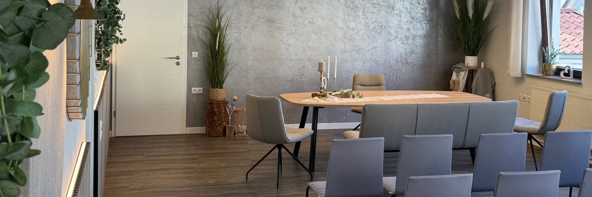 Modern eingerichteter Trau- und Veranstaltungsraum mit großem Holztisch, grauen Stühlen und Sitzbank. Über dem Tisch hängen zwei runde Leuchten; Kerzen und florale Dekoration schmücken die Tischmitte. An den Wänden befinden sich Pflanzen, runde Akustikelemente und eine graue Strukturwand.