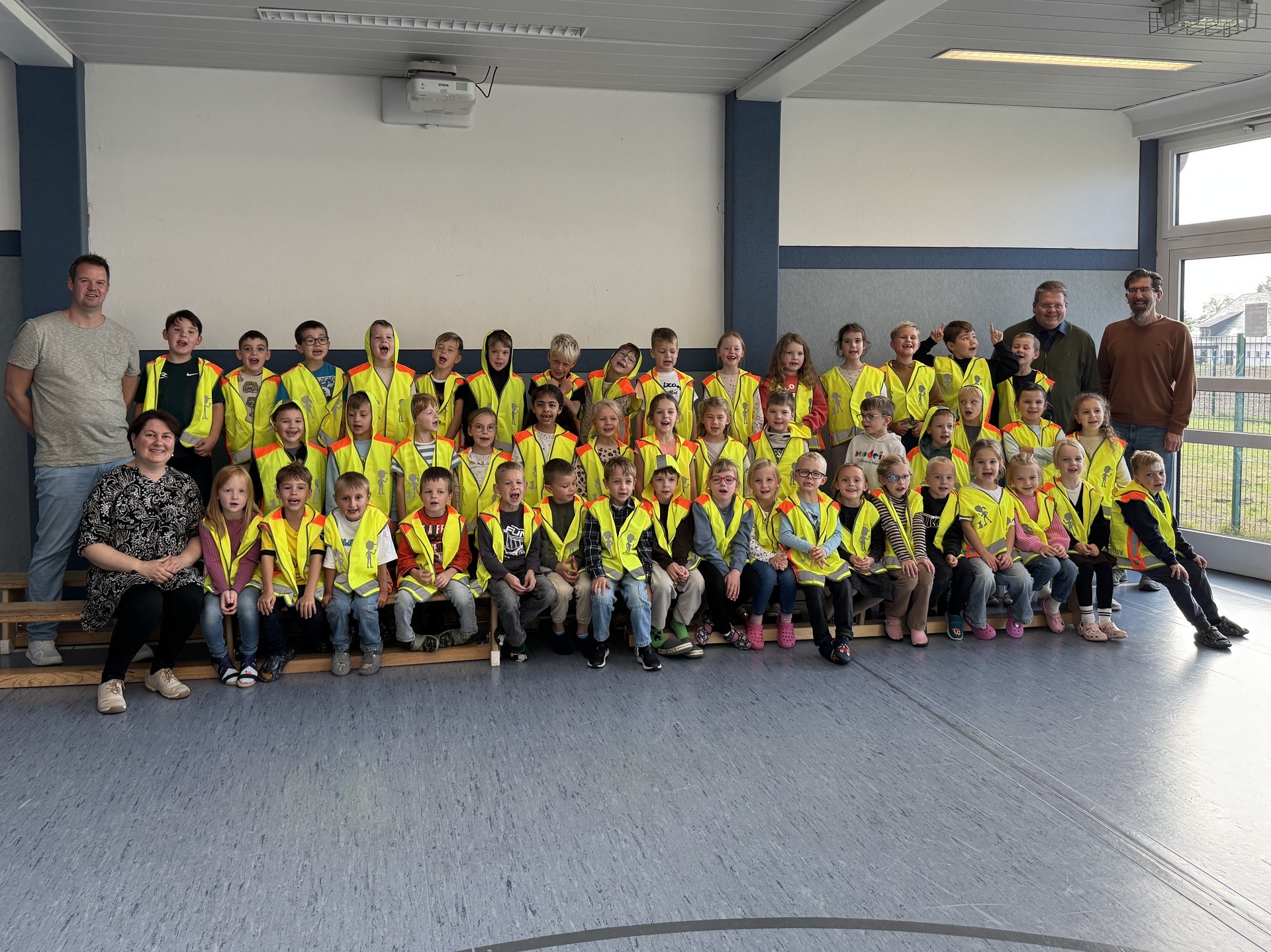 Gruppenfoto in einer Schule: Rund 40 Kinder tragen leuchtend gelbe Warnwesten und sitzen oder stehen in drei Reihen in einem hellen Raum. Links und rechts begleiten vier erwachsene Personen die Gruppe. Im Hintergrund sind eine weiße Wand, blaue Säulen und große Fenster zu sehen.