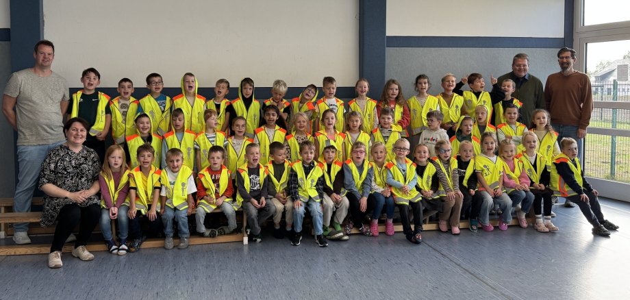 Gruppenfoto in einer Schule: Rund 40 Kinder tragen leuchtend gelbe Warnwesten und sitzen oder stehen in drei Reihen in einem hellen Raum. Links und rechts begleiten vier erwachsene Personen die Gruppe. Im Hintergrund sind eine weiße Wand, blaue Säulen und große Fenster zu sehen.