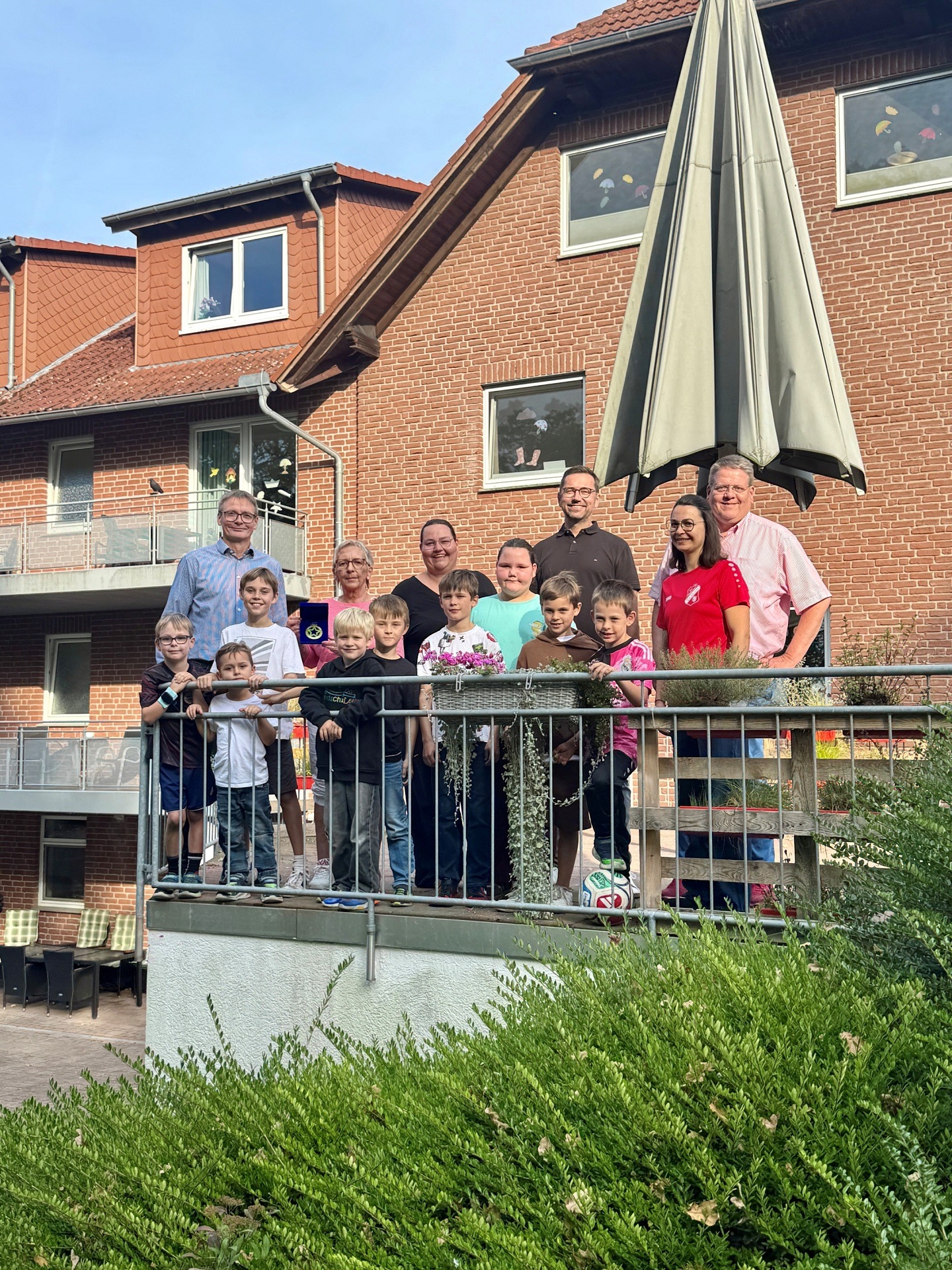 Eine Gruppe aus Erwachsenen und Kindern steht bei sonnigem Wetter hinter einem Metallgeländer vor einem roten Klinkergebäude mit Balkonen und Dachgauben. Im Hintergrund ist ein großer geschlossener Sonnenschirm zu sehen. Ein Junge hält einen Fußball in den Händen, einige Kinder tragen Sportkleidung.