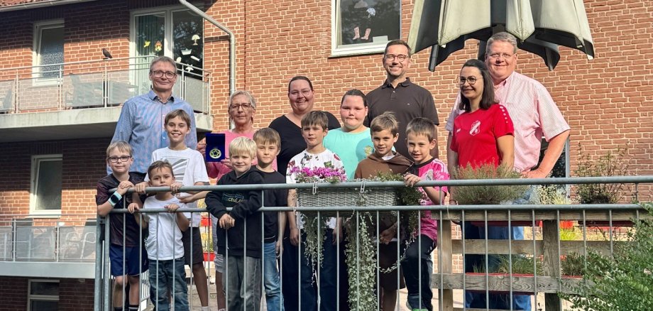 LokalStars 2026 Eine Gruppe aus Erwachsenen und Kindern steht bei sonnigem Wetter hinter einem Metallgeländer vor einem roten Klinkergebäude mit Balkonen und Dachgauben. Im Hintergrund ist ein großer geschlossener Sonnenschirm zu sehen. Ein Junge hält einen Fußball in den Händen, einige Kinder tragen Sportkleidung.