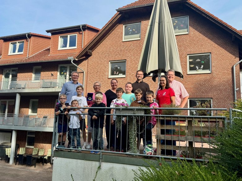 LokalStars 2026 Eine Gruppe aus Erwachsenen und Kindern steht bei sonnigem Wetter hinter einem Metallgeländer vor einem roten Klinkergebäude mit Balkonen und Dachgauben. Im Hintergrund ist ein großer geschlossener Sonnenschirm zu sehen. Ein Junge hält einen Fußball in den Händen, einige Kinder tragen Sportkleidung.
