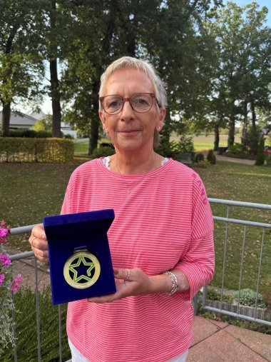 LokalStars 2026 Eine Frau in einem rosa gestreiften Oberteil steht im Freien vor Bäumen und einem Garten. Sie hält eine geöffnete dunkelblaue Schatulle mit der Medaille „LokalStars 2026 – Der Award für nachhaltiges Engagement“ in die Kamera.