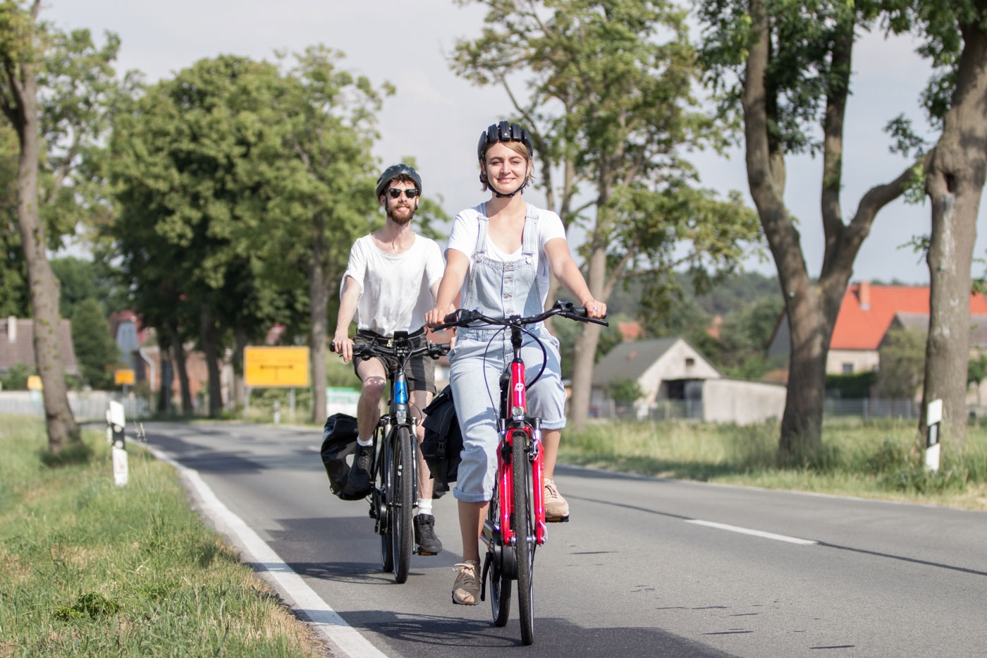Zwei Radfahrende mit Fahrradhelmen fahren auf einer Landstraße durch eine grüne, von Bäumen gesäumte Landschaft. Im Vordergrund fährt eine Frau auf einem roten Fahrrad, hinter ihr ein Mann auf einem schwarzen Fahrrad.