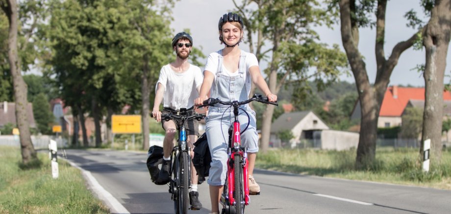 Fahrradklima-Test 2026 Zwei Radfahrende mit Fahrradhelmen fahren auf einer Landstraße durch eine grüne, von Bäumen gesäumte Landschaft. Im Vordergrund fährt eine Frau auf einem roten Fahrrad, hinter ihr ein Mann auf einem schwarzen Fahrrad.