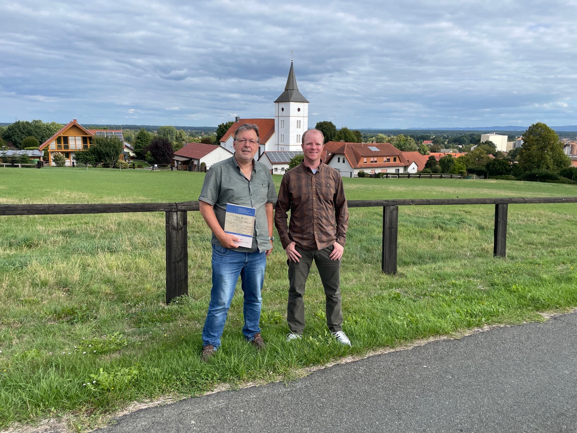 Zwei Männer stehen vor einem Holzzaun auf einer grünen Wiese. Im Hintergrund sind Häuser, eine Kirche mit Turm und eine weite Landschaft unter einem bewölkten Himmel zu sehen. Der Mann links hält eine Mappe in der Hand.