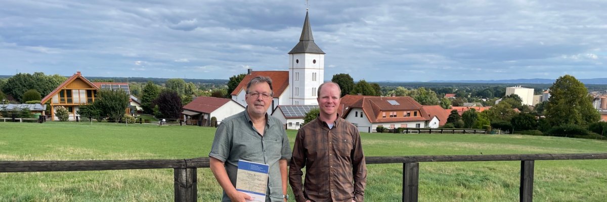 Vorfahrensuche 2026 Zwei Männer stehen vor einem Holzzaun auf einer grünen Wiese. Im Hintergrund sind Häuser, eine Kirche mit Turm und eine weite Landschaft unter einem bewölkten Himmel zu sehen. Der Mann links hält eine Mappe in der Hand.