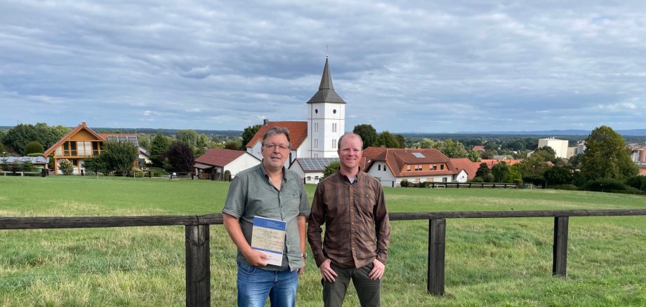 Vorfahrensuche 2026 Zwei Männer stehen vor einem Holzzaun auf einer grünen Wiese. Im Hintergrund sind Häuser, eine Kirche mit Turm und eine weite Landschaft unter einem bewölkten Himmel zu sehen. Der Mann links hält eine Mappe in der Hand.