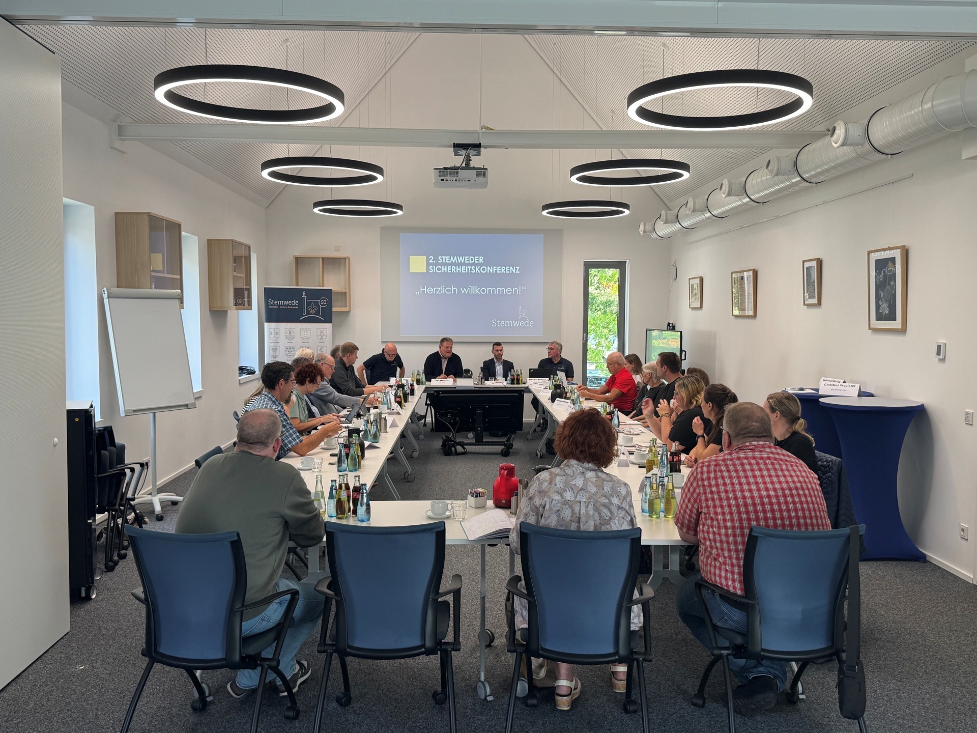 Etwa 20 Personen sitzen an langen Tischen in einem hellen Sitzungsraum. Vorne sitzen mehrere Männer an einem Podium; auf der Leinwand steht „2. Stemweder Sicherheitskonferenz – Herzlich willkommen!“.