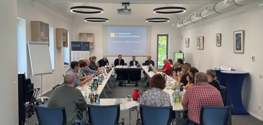 Stemweder Sicherheitskonferenz 2026 Etwa 20 Personen sitzen an langen Tischen in einem hellen Sitzungsraum. Vorne sitzen mehrere Männer an einem Podium; auf der Leinwand steht „2. Stemweder Sicherheitskonferenz – Herzlich willkommen!“.