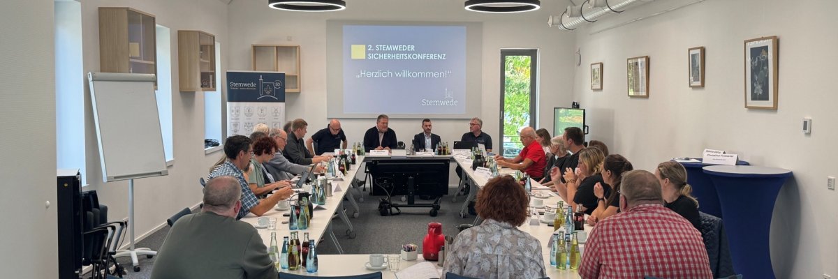 Stemweder Sicherheitskonferenz 2026 Etwa 20 Personen sitzen an langen Tischen in einem hellen Sitzungsraum. Vorne sitzen mehrere Männer an einem Podium; auf der Leinwand steht „2. Stemweder Sicherheitskonferenz – Herzlich willkommen!“.