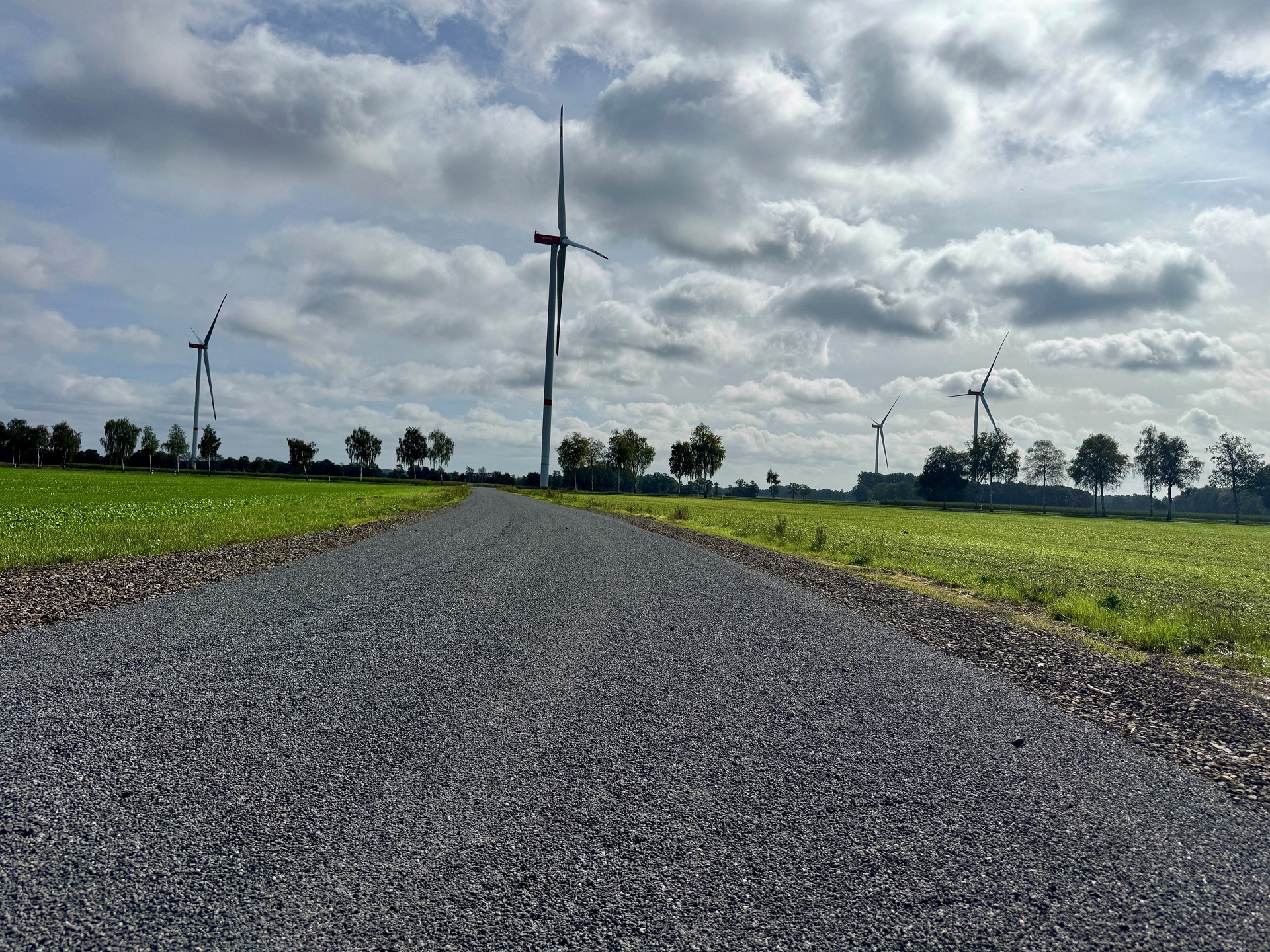 Asphaltierter Wirtschaftsweg führt durch grüne Felder zu mehreren Windenergieanlagen unter einem wolkigen Himmel.