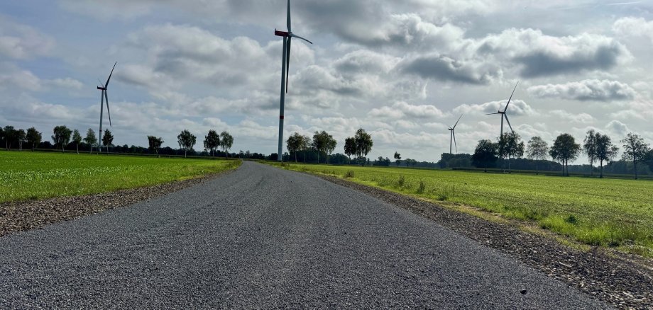Wege Tiefenriede 2026 Asphaltierter Wirtschaftsweg führt durch grüne Felder zu mehreren Windenergieanlagen unter einem wolkigen Himmel.