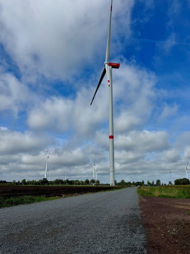 Wege Tiefenriede 2026 Große Windenergieanlage mit roten Markierungen steht neben einem Wirtschaftsweg inmitten von Feldern; weitere Anlagen sind im Hintergrund unter blauem, wolkigem Himmel zu sehen.