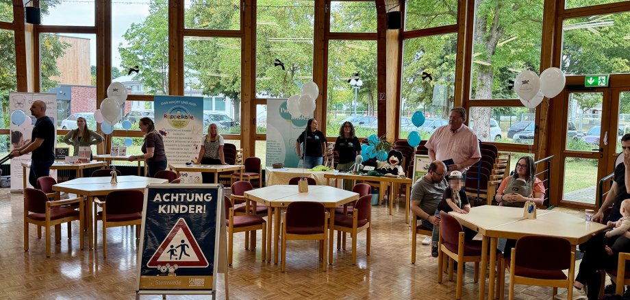 Neugeborenenbegrüßung In einem hellen Veranstaltungsraum mit großen Fenstern informieren verschiedene Stände junge Familien. Mehrere Erwachsene sitzen mit kleinen Kindern an Tischen, während im Hintergrund Informationsmaterial, Luftballons und ein Schild mit der Aufschrift „Achtung Kinder!“ zu sehen sind.