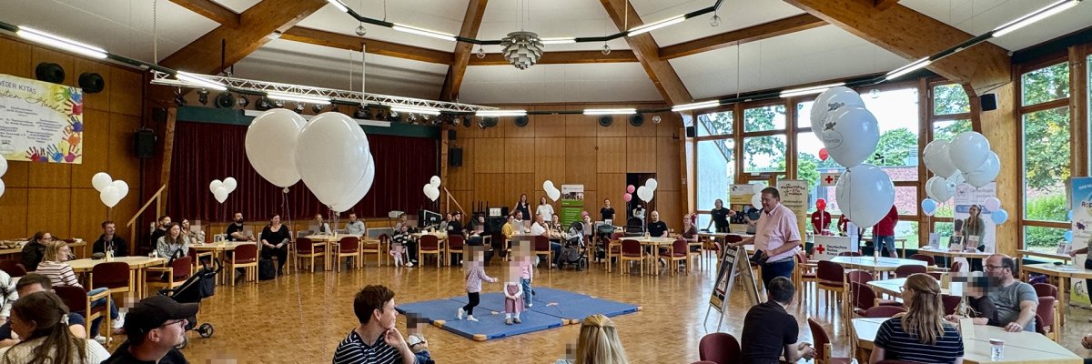 Neugeborenenbegrüßung 2026 In einem großen, hellen Saal sitzen junge Familien an Tischen. Kinder spielen auf Matten in der Mitte des Raumes. Informationsstände und weiße Luftballons sind rundherum aufgebaut; Bürgermeister Kai Abruszat begrüßt die Gäste.