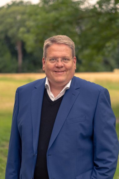 Ein Portraitfoto eines Mannes mit hellbraunen, kurzen Haaren, einer Brille, der ein blaues Jacket und darunter einen schwarzen Pullover trägt. Er steht vor einem Kornfeld, das im Hintergrund verschwommen zu erkennen ist.