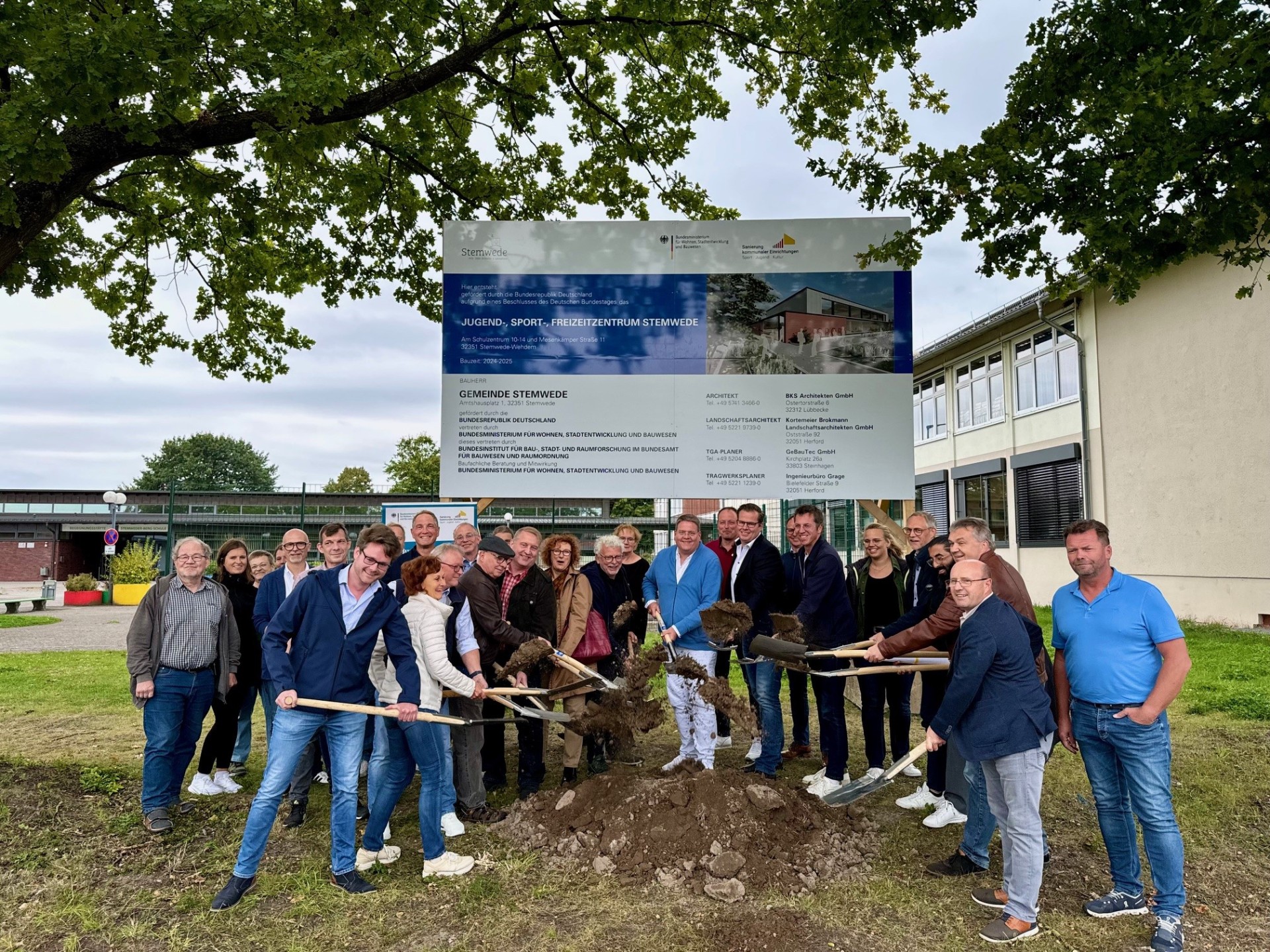 Rund 20 Personen, die unter einem großen Baustellenschild stehen. Einige Akteure haben eine Spaten in der Hand und werfen damit etwas Erde in die Höhe, als Zeichen des symbolischen Spatenstichs zum Projekt.