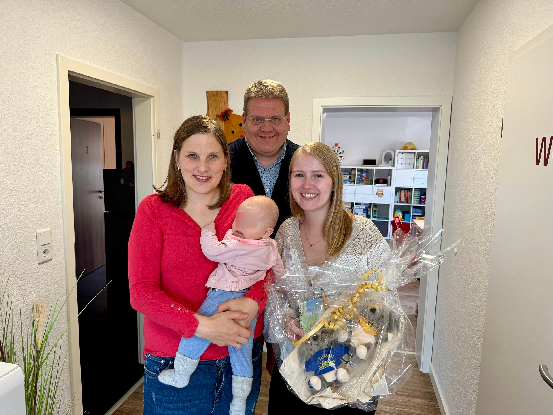 Zwei junge Frauen - die eine trägt ein Baby auf dem Arm, die andere hält ein Geschenk in den Händen - lächeln in die Kamera. Im Hintergrund steht Stemwedes Bürgermeister.