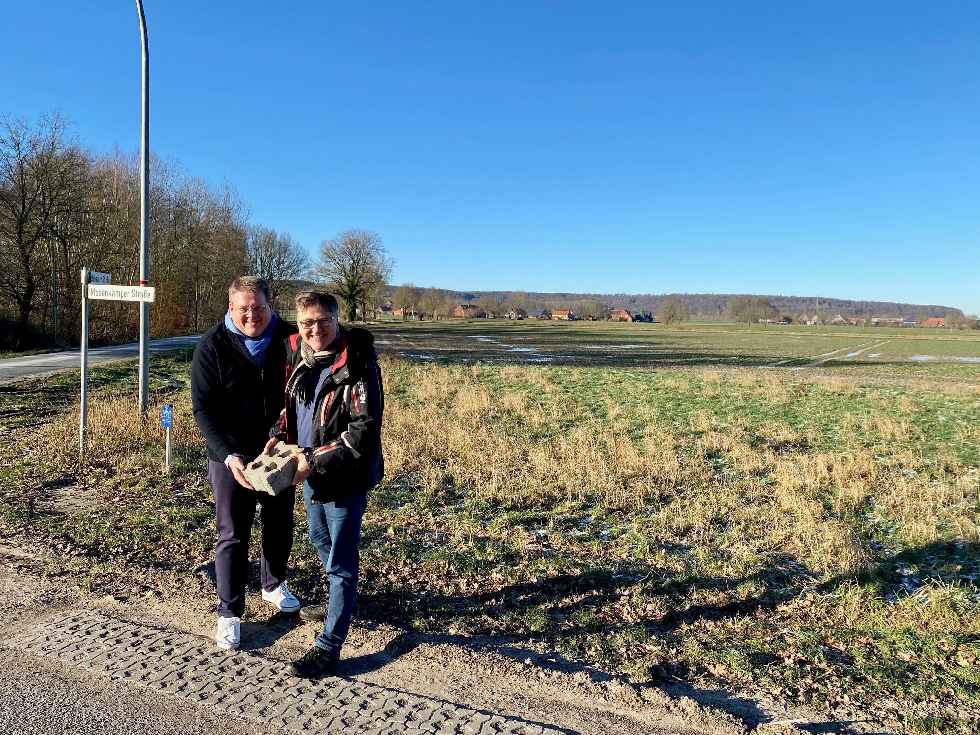 Zwei Männer stehen an einer Straße und halten exemplarisch einen Rasengitterstein, der am Wegesrand verbaut wird. 