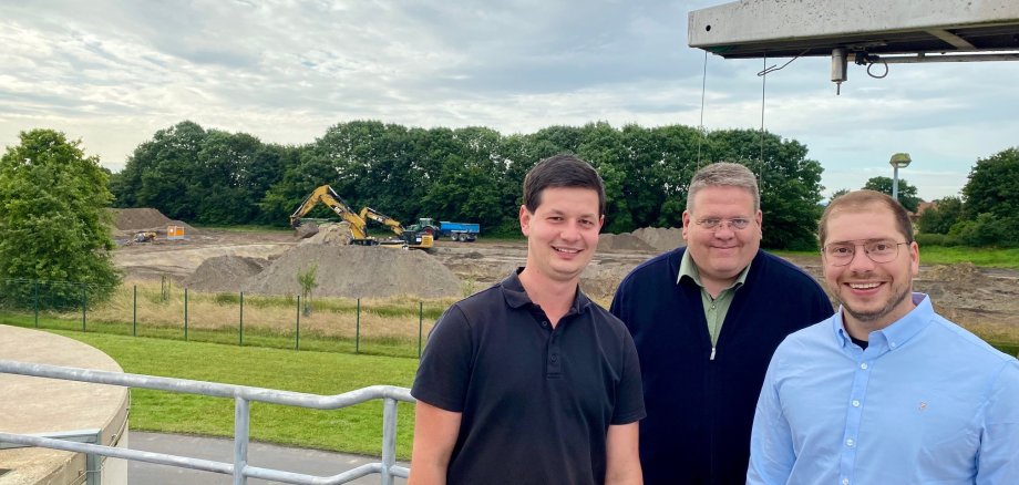 Drei Männer, die vor einer ausgebaggerten Fläche einer großen Baustelle stehen und in die Kamera lächeln.