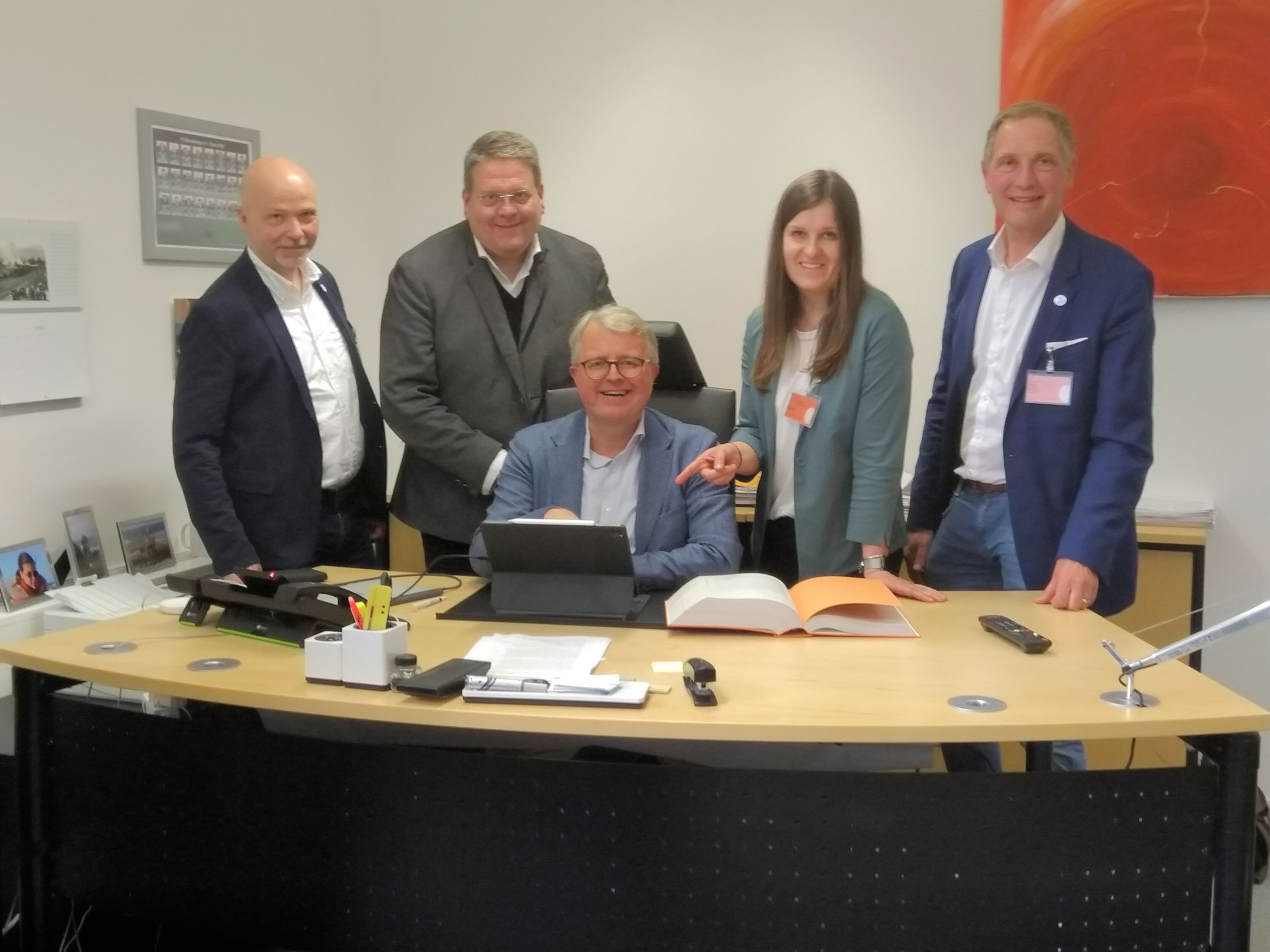 Ein Mann sitzt an einem Laptop an einem Schreibtisch. Hinter ihm stehen drei Männer und eine Frau uns lächeln in die Kamera.