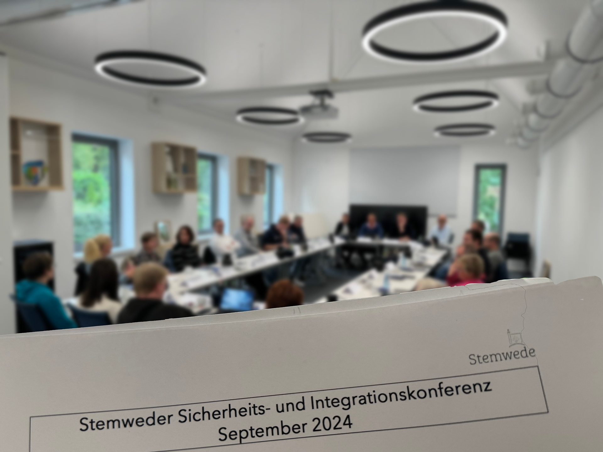 Ein nur unscharf zu erkennender Sitzungssaal in dem zahlreiche Personen sitzen. Im Vordergrund ist ein Papier zu sehen auf dem folgendes steht: Stemweder Sicherheits- und Integrationskonferenz, September 2024".