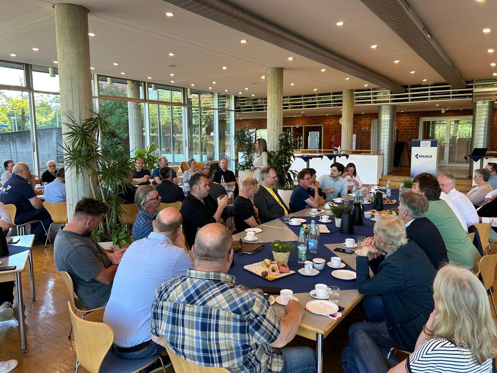 Etwa 50 Personen, die in einer Betriebskantine an verschiedenen, eingedeckten Frühstückstafeln sitzen und einer Person, die im Hintergrund steht, zuhören.