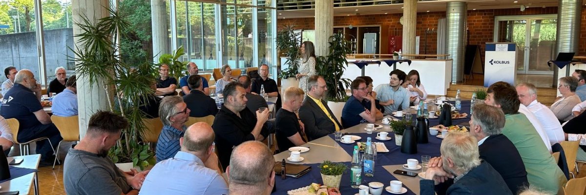 Etwa 50 Personen, die in einer Betriebskantine an verschiedenen, eingedeckten Frühstückstafeln sitzen und einer Person, die im Hintergrund steht, zuhören.