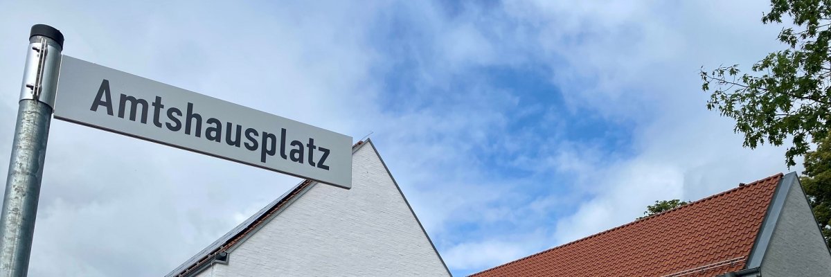 Ein Straßenschild, mit der Aufschrift "Amtshausplatz" vor einem modernen, weißen Gebäude, an dem die Aufschrift "Amtshaus" zu lesen ist.