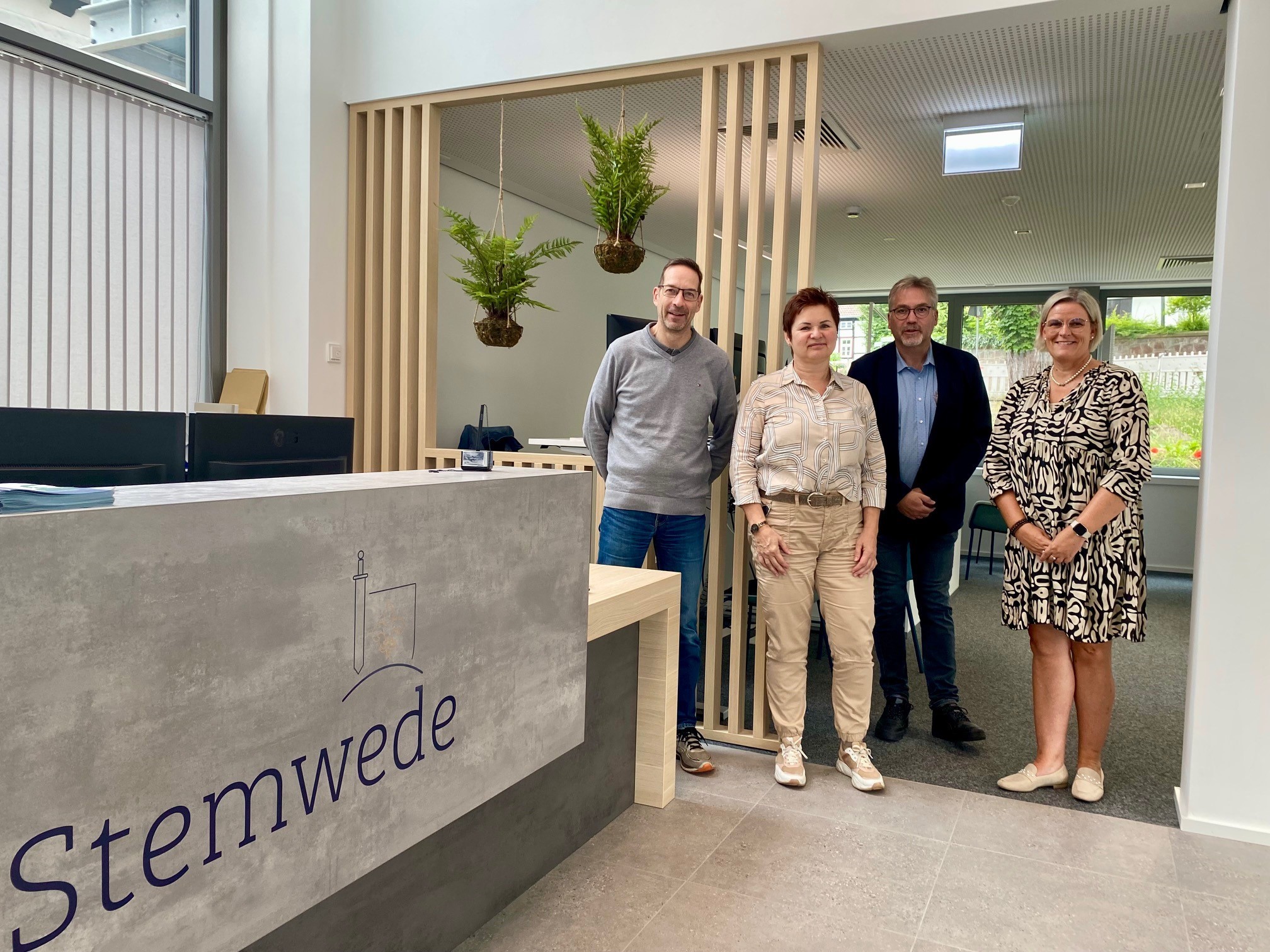Zwei Männer und zwei Frauen stehen in einem modernen Eingangsbereich einer Verwaltung. Auf dem Empfangstresen im linken Teil des Fotos, ist das Logo der Gemeinde Stemwede zu erkennen.