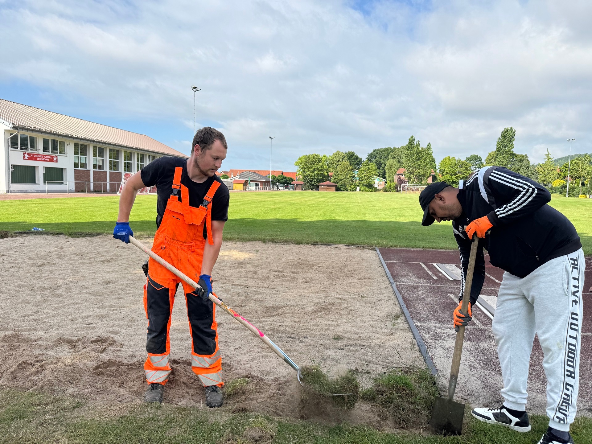 Zwei Männer arbeiten an einer Weitsprunggrube; einer trägt orange Arbeitskleidung und harkt Sand, der andere trägt Sportkleidung und gräbt am Rand mit einer Schaufel.